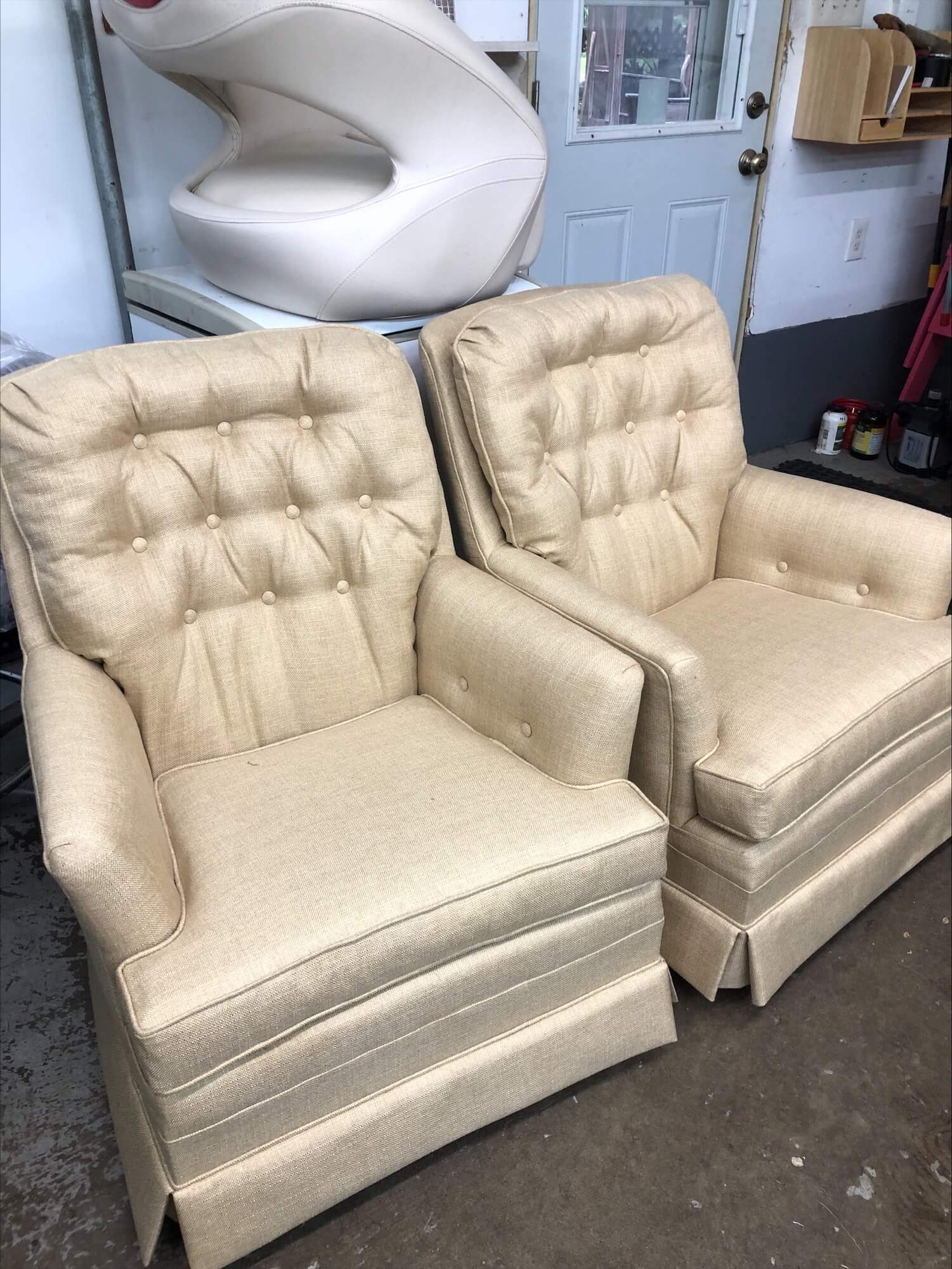 Two beige, tufted armchairs with loose cushions are positioned in a garage space, showcasing their classic design and neutral fabric.