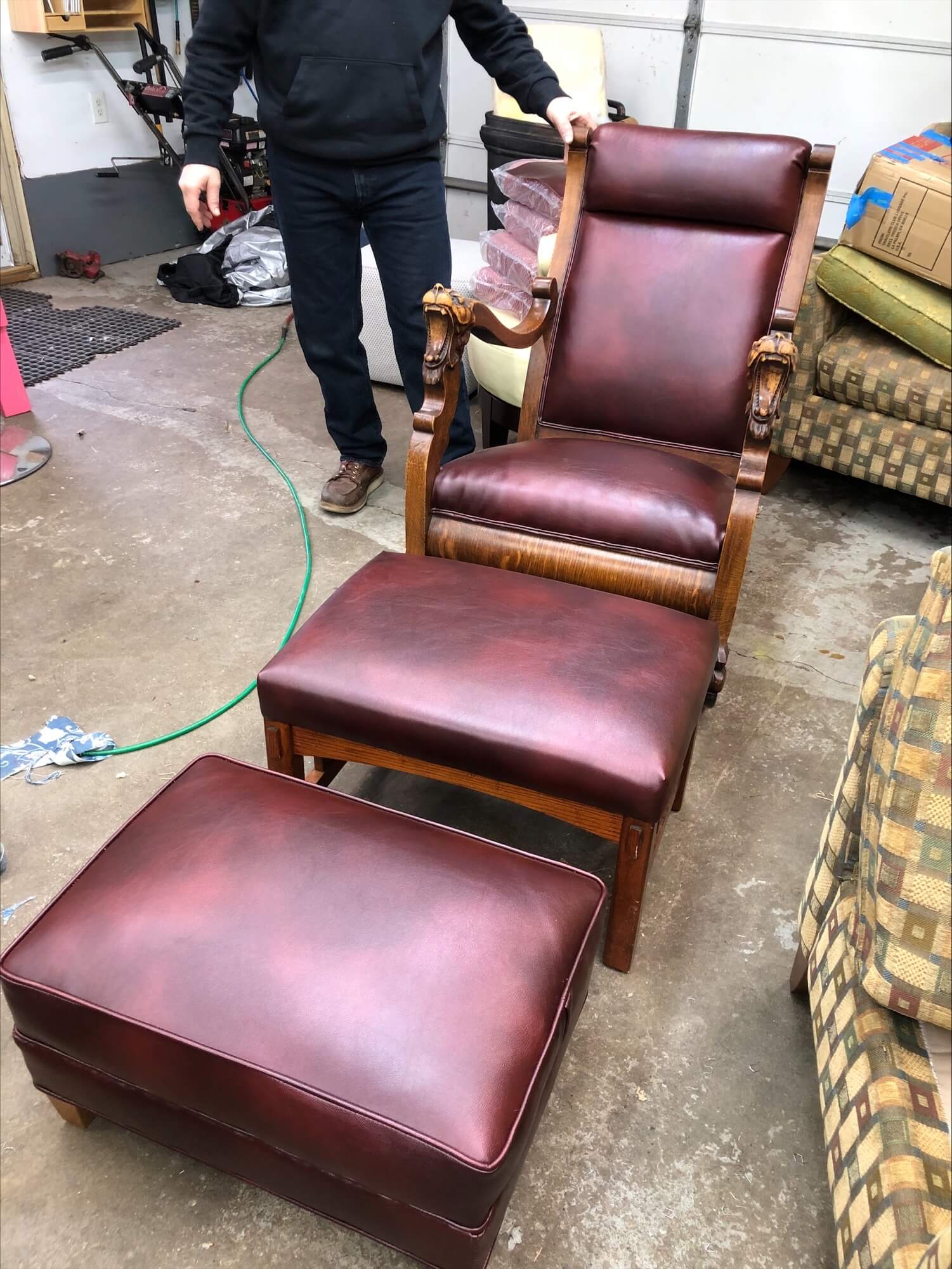 A wooden chair with dark red leather upholstery and ornate armrest carvings, accompanied by a matching ottoman, is displayed in a garage setting.