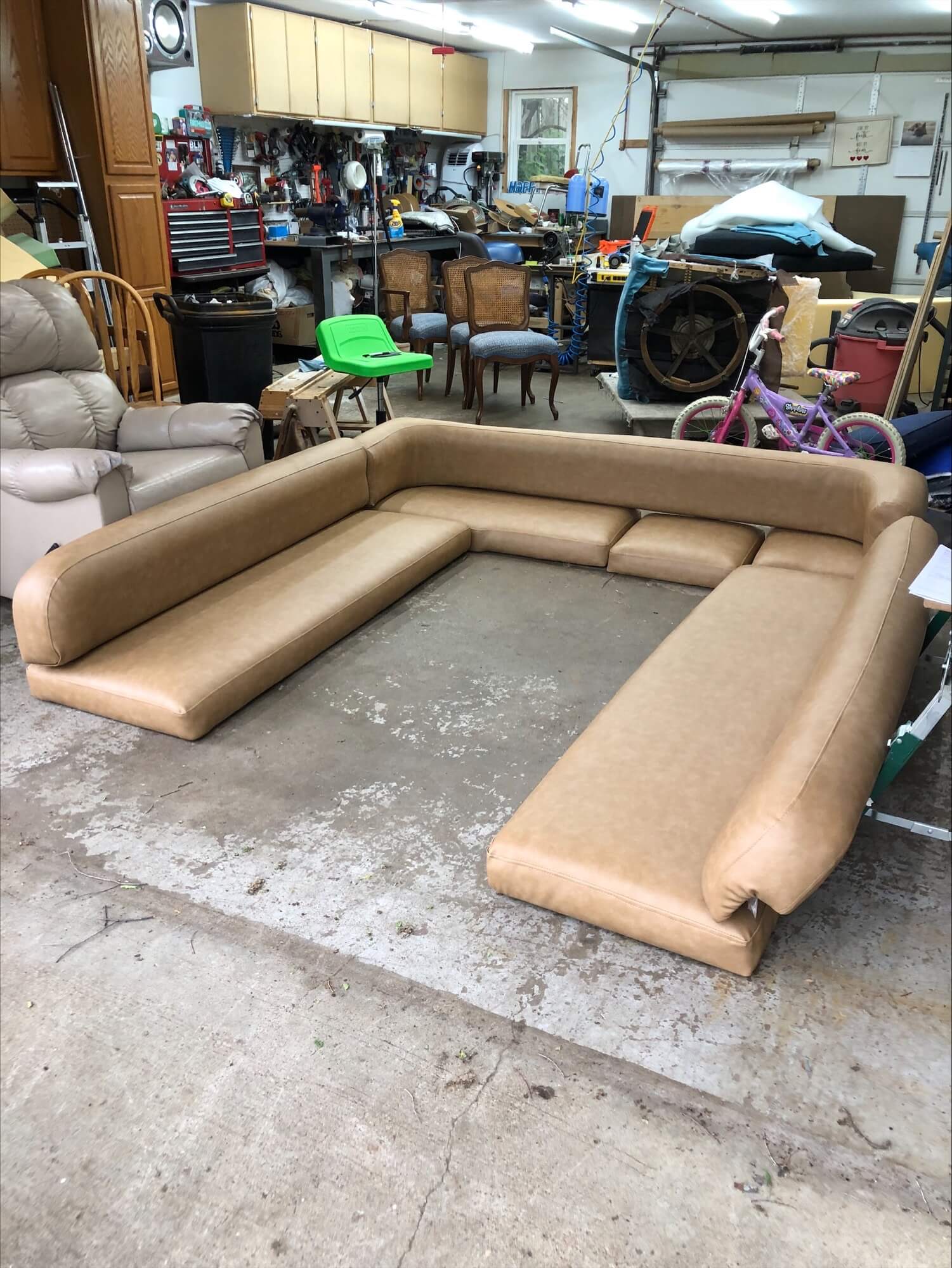 A large, minimalist tan sectional sofa is arranged in a cluttered garage, surrounded by tools, chairs, and various storage items, highlighting its casual setting.